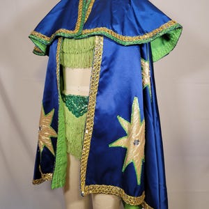 May include: A blue cape with gold star accents and gold trim. The cape has a green lining and is paired with a green fringed top. The costume is displayed on a mannequin.