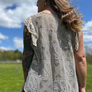 Puede incluir: Un top blanco de crochet con un patrón geométrico. El top tiene un escote redondo y mangas cortas con un borde de volantes.