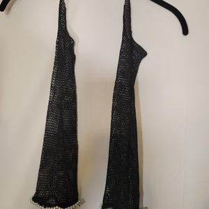 May include: Black mesh arm warmers with a halter neck design. The arm warmers are adorned with a row of small, white pearls at the wrist. The arm warmers are hanging on a black hanger against a white background.