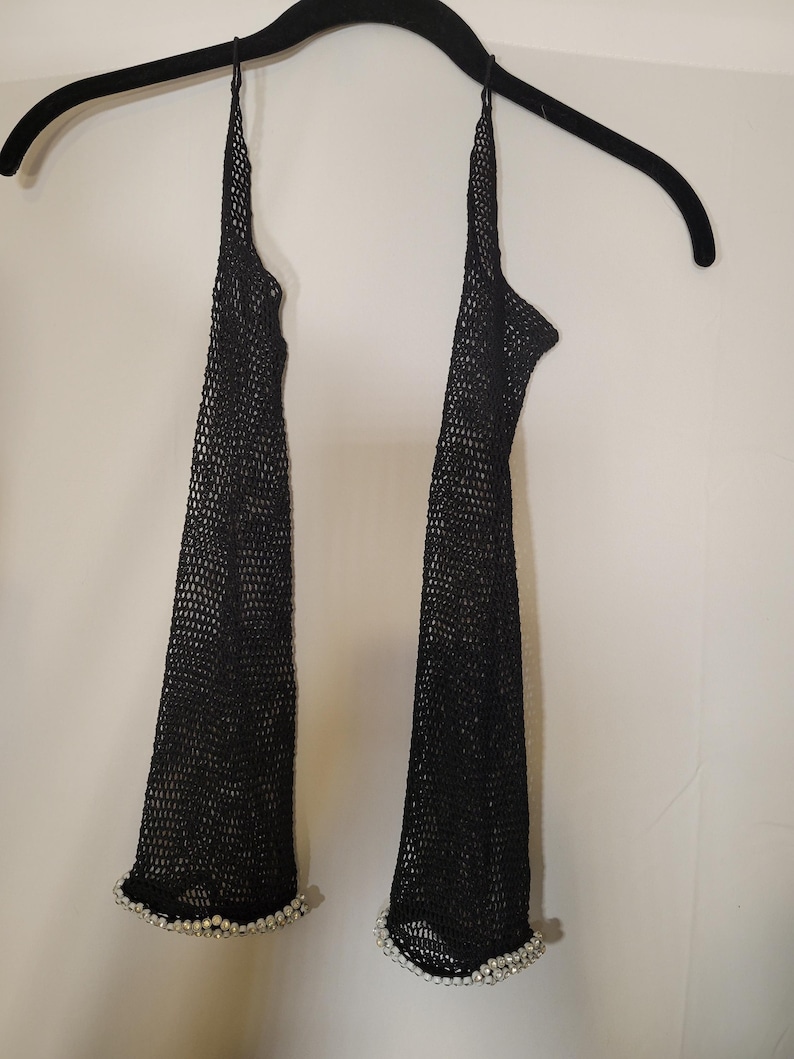 May include: Black mesh arm warmers with a halter neck design. The arm warmers are adorned with a row of small, white pearls at the wrist. The arm warmers are hanging on a black hanger against a white background.