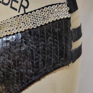 May include: Black sequined garment with a decorative band of white and gold-toned embellishments. The garment has black straps and is displayed on a mannequin.