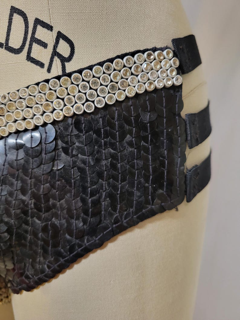May include: Black sequined garment with a decorative band of white and gold-toned embellishments. The garment has black straps and is displayed on a mannequin.