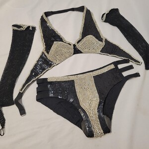 May include: A black bikini set with silver embellishments. The set includes a halter top, high-waisted bottoms, and two arm sleeves. The bikini top and bottoms have a sequined design with silver accents. The arm sleeves are made of a mesh material.