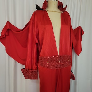 Könnte beinhalten: Ein rotes Teufelskostüm bestehend aus einem Satin-Jumpsuit mit tiefem V-Ausschnitt, einem breiten Pailletten-Taillenband und passenden Pailletten-Manschetten. Das Outfit umfasst einen an den Ärmeln befestigten Umhang, einen Stehkragen und ein Stirnband.