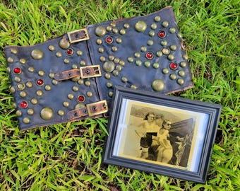 May include: Black leather corset with brass studs and red gems, featuring three buckle closures. A framed black and white photograph of three people is next to the corset, resting on green grass.