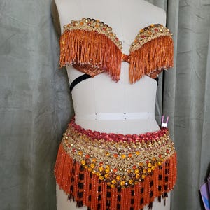 Puede incluir: Un conjunto de disfraz con cuentas de color naranja y dorado, que incluye un sujetador y una parte inferior similar a una falda. El sujetador presenta flecos y abalorios decorativos. La falda tiene una parte superior con cuentas y flecos. El disfraz se exhibe en un maniquí.