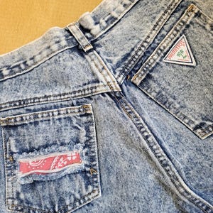 May include: Close-up of acid wash denim jeans. The jeans feature a back pocket with a pink paisley patch and a triangular label that reads "Palmettos". The denim has a faded, worn look.
