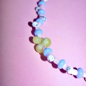 Könnte beinhalten: Eine Perlenkette mit einem sich wiederholenden Muster aus hellblauen Scheibenperlen, kleinen transparenten rosa Zwischenperlen und weißen Perlen mit schwarzen Sprenkeln. Eine größere, glatte, blassgelbe, kleeblattförmige Perle bildet den Mittelpunkt.