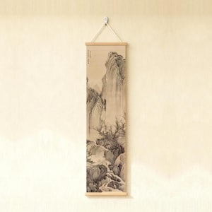 May include: A vertical wall hanging scroll featuring a monochromatic ink wash landscape painting of towering mountains, rocky terrain, and bare trees. The artwork is mounted on light wood rods and suspended by a white cord.