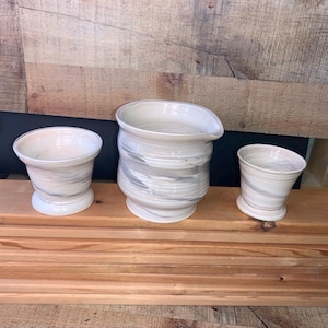 May include: Three handmade ceramic pieces: a small cup, a pitcher with a spout, and a slightly angled cup. All are white with gray swirling patterns. The pieces are displayed on a wooden surface against a rustic wood background.