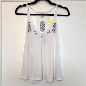 May include: White camisole top with a V-neck and spaghetti straps, hanging on a black hanger. The top features embroidered floral designs in gray and black. A yellow tag is attached to the strap.
