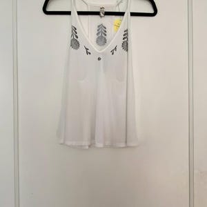 May include: White tank top with a V-neck and floral embroidery. The sleeveless top has a loose fit and thin straps. The floral design is in a dark gray color. The top is hanging on a black hanger.