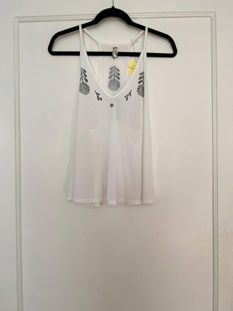 May include: White tank top with a V-neck and floral embroidery. The sleeveless top has a loose fit and thin straps. The floral design is in a dark gray color. The top is hanging on a black hanger.