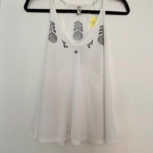 May include: White camisole top with a V-neck and floral embroidery. The sleeveless top is hanging on a black hanger. The embroidery is black and is on the front of the top.
