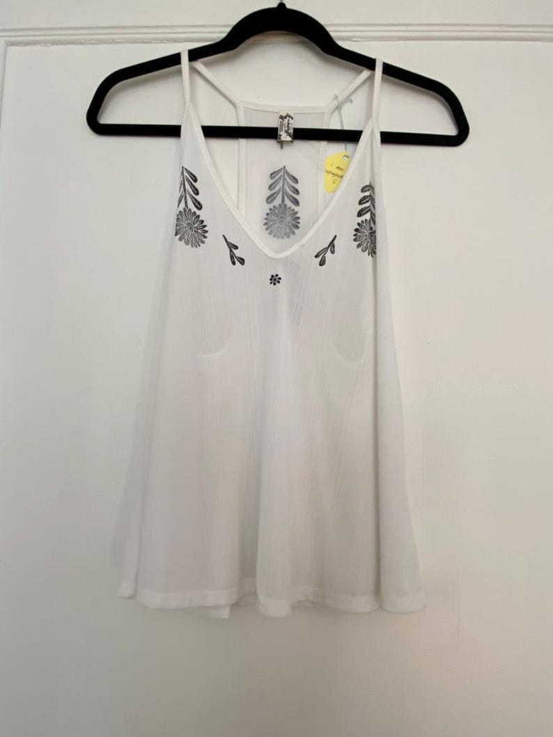 May include: White camisole top with a V-neck and floral embroidery. The sleeveless top is hanging on a black hanger. The embroidery is black and is on the front of the top.