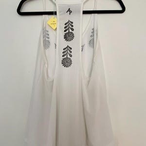 May include: White tank top with a floral design on the back. The tank top is hanging on a black hanger. The back of the tank top has a vertical floral pattern and the letter "A".