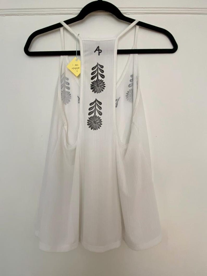 May include: White tank top with a floral design on the back. The tank top is hanging on a black hanger. The back of the tank top has a vertical floral pattern and the letter "A".