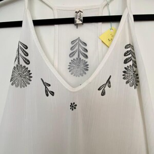May include: White camisole with a V-neck and spaghetti straps. The camisole features black floral and leaf patterns. A small tag is attached to the garment. The camisole is hanging on a black hanger.