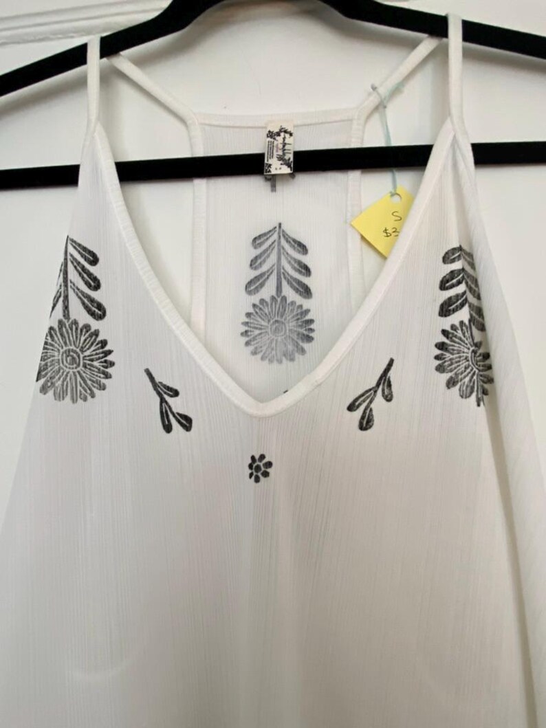 May include: White camisole with a V-neck and spaghetti straps. The camisole features black floral and leaf patterns. A small tag is attached to the garment. The camisole is hanging on a black hanger.