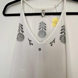 May include: White camisole with a V-neck and thin straps, adorned with black floral and leaf patterns. The top is hanging on a black hanger. A yellow tag reads "I am upcycled".