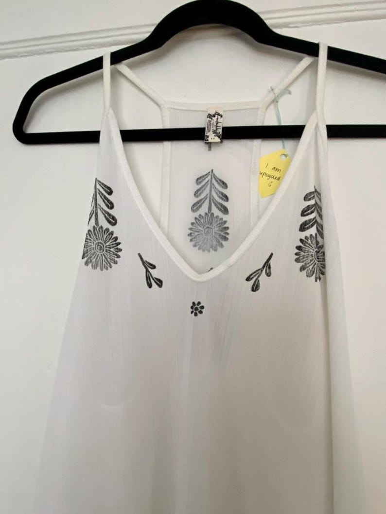 May include: White camisole with a V-neck and thin straps, adorned with black floral and leaf patterns. The top is hanging on a black hanger. A yellow tag reads "I am upcycled".