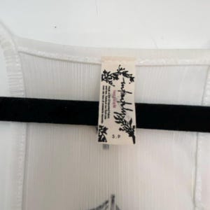 May include: White sleeveless top with a black graphic design and a small tag. The top is hanging on a black hanger. The tag has the words "free people" and "S.P."