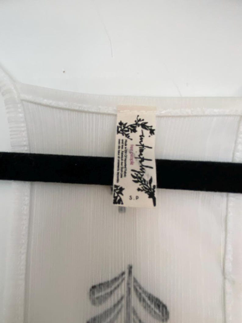 May include: White sleeveless top with a black graphic design and a small tag. The top is hanging on a black hanger. The tag has the words "free people" and "S.P."