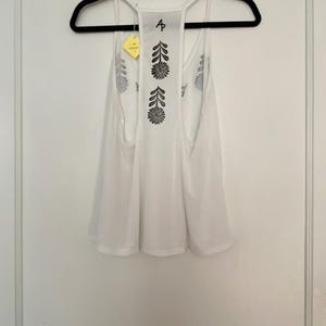 May include: White tank top with a floral design on the back, hanging on a black hanger. The tank top has a racerback style and a loose fit. The floral design is black and appears to be screen printed. A yellow tag is attached to the hanger.