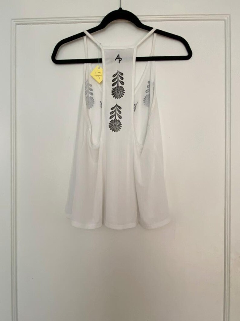 May include: White tank top with a floral design on the back, hanging on a black hanger. The tank top has a racerback style and a loose fit. The floral design is black and appears to be screen printed. A yellow tag is attached to the hanger.