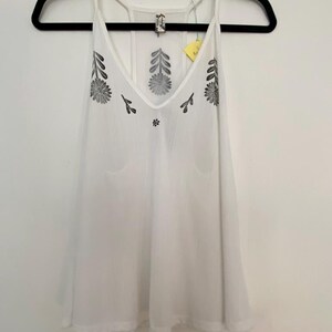 May include: White camisole top with a V-neck and spaghetti straps. The top features black floral embroidery around the neckline and on the front. The garment is hanging on a black hanger against a white wall.