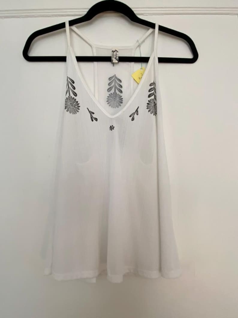 May include: White camisole top with a V-neck and spaghetti straps. The top features black floral embroidery around the neckline and on the front. The garment is hanging on a black hanger against a white wall.