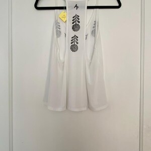 May include: White tank top with a floral design in black ink. The tank top is hanging on a black hanger. The back of the tank top has a floral design. A yellow tag is attached to the tank top.