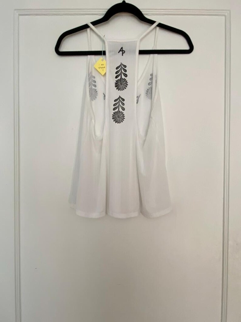 May include: White tank top with a floral design in black ink. The tank top is hanging on a black hanger. The back of the tank top has a floral design. A yellow tag is attached to the tank top.