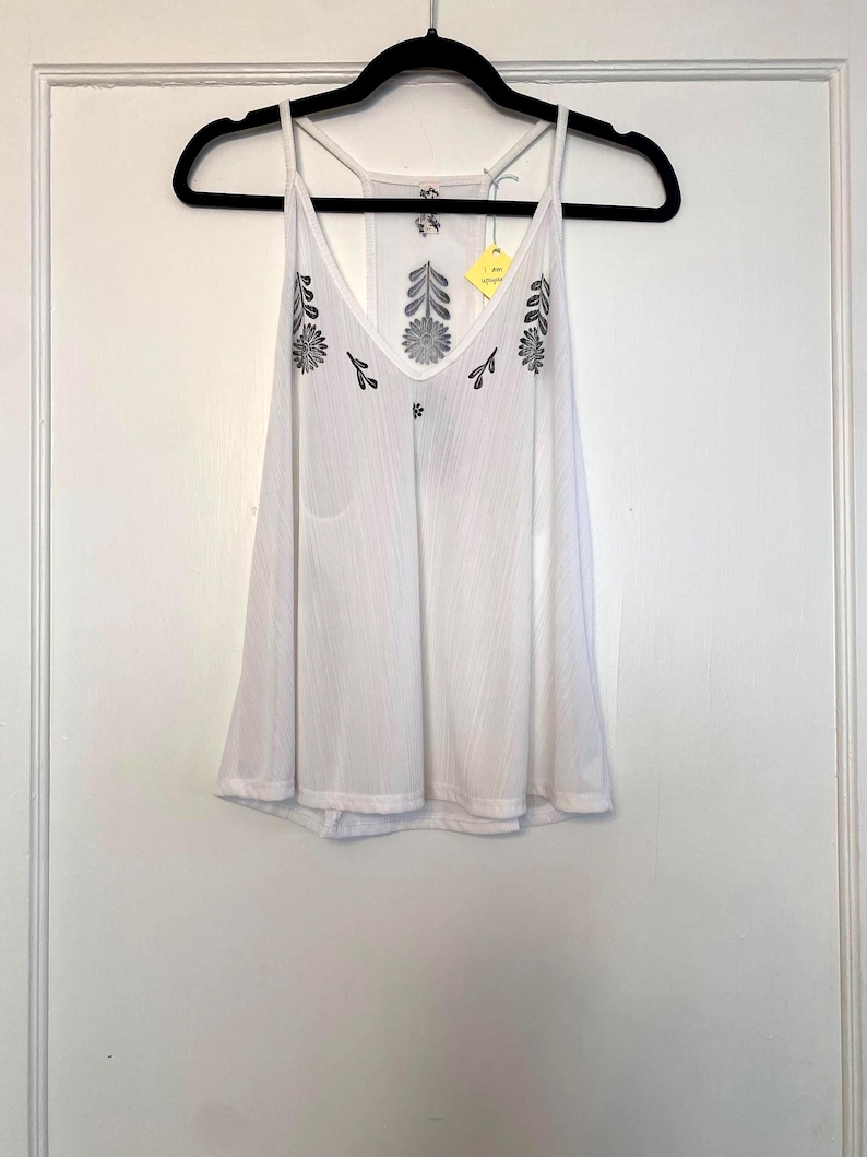 May include: White camisole top with a V-neck and spaghetti straps, hanging on a black hanger. The top features embroidered floral designs in gray and black. A yellow tag is attached to the strap.