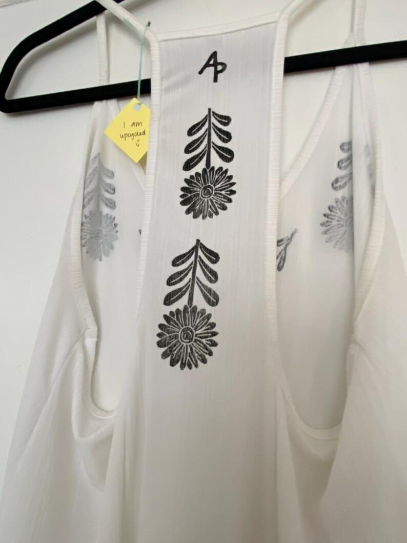 May include: White tank top with black floral and leaf print. The back features a central panel with two large flower designs and leaf accents. The top has a tag that says "I am upcycled".