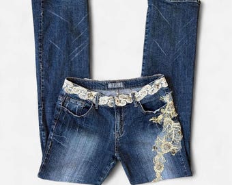 May include: Dark blue denim jeans featuring a straight-leg cut with whiskering details. The waistband is adorned with a continuous band of white and gold floral lace embroidery, which extends down the left hip in a large, intricate floral pattern.