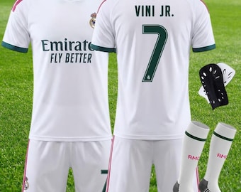 May include: A white soccer jersey and shorts set featuring dark green and pink accents. The jersey displays the text Emirates FLY BETTER, the Real Madrid crest, and the name VINI JR. with the number 7 on the back. Includes black shin guards and white socks.
