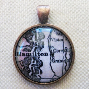 May include: A round, copper-toned pendant with a glass insert featuring a vintage map of Hamilton, Ontario, Canada. The map shows the surrounding towns of Victor, Corvallis, and Grantsdale.