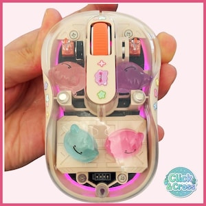 Puede incluir: Un ratón de ordenador inalámbrico con carcasa transparente, que presenta una rueda de desplazamiento naranja e iluminación LED rosa interna. El dispositivo contiene cuatro pequeñas figuras translúcidas con forma de criatura y el logo Click & Cross.