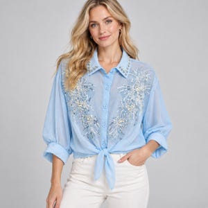Puede incluir: Una camisa azul claro de botones con mangas largas, cuello y un dobladillo frontal para anudar. La camisa está decorada con intrincados bordados florales blancos y plateados, lentejuelas y cuentas tipo perla en los paneles frontales y el cuello.