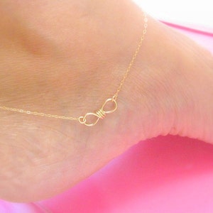 May include: A delicate gold-coloured anklet featuring an infinity symbol. The anklet is worn on a foot, with the infinity symbol resting on the top of the foot. The chain is thin and the infinity symbol is a wire design.