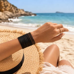 May include: A stack of thin black bangles worn on the wrist, accompanied by a gold ring with a small white stone on the finger. The jewelry is displayed against a straw hat with a black band, featuring a textured weave and frayed edges.