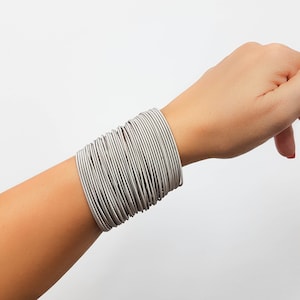 May include: A stack of silver bangles worn on a person's wrist. The bangles are thin and create a thick band around the wrist.