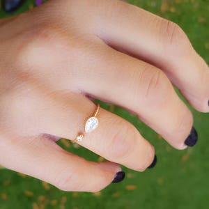 Op de afbeelding: Een gouden ring met een traanvormige, heldere steen en een kleinere heldere steen op de band.