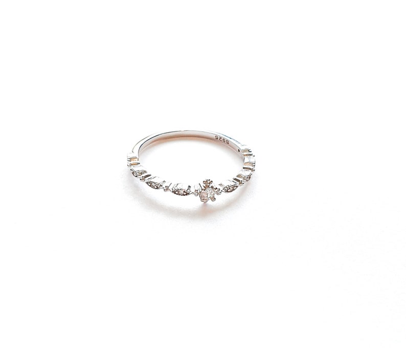 May include: A silver ring with a small, clear stone in the center and smaller stones set around the band.