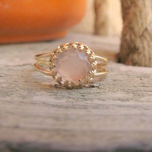 Op de afbeelding: Een delicate gouden ring met een lichtroze edelsteen gezet in een decoratieve, gegolfde bezel. De ring heeft een dubbele band.