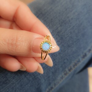 Op de afbeelding: Een gouden ring met een blauwe opaal-steen gezet in een delicaat, versierd frame. De ring wordt gedragen aan een vinger.