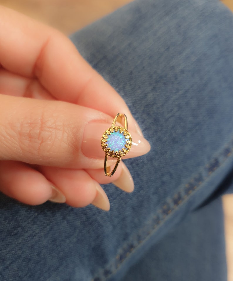 Op de afbeelding: Een gouden ring met een blauwe opaal-steen gezet in een delicaat, versierd frame. De ring wordt gedragen aan een vinger.