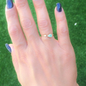 Op de afbeelding: Een gouden ring met een traanvormige turquoise steen en een kleine ronde, heldere steen. De ring zit aan een vinger.