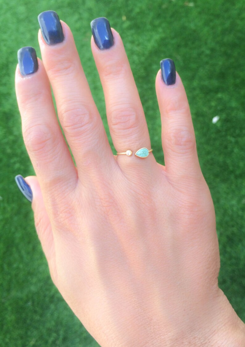 Op de afbeelding: Een gouden ring met een traanvormige turquoise steen en een kleine ronde, heldere steen. De ring zit aan een vinger.